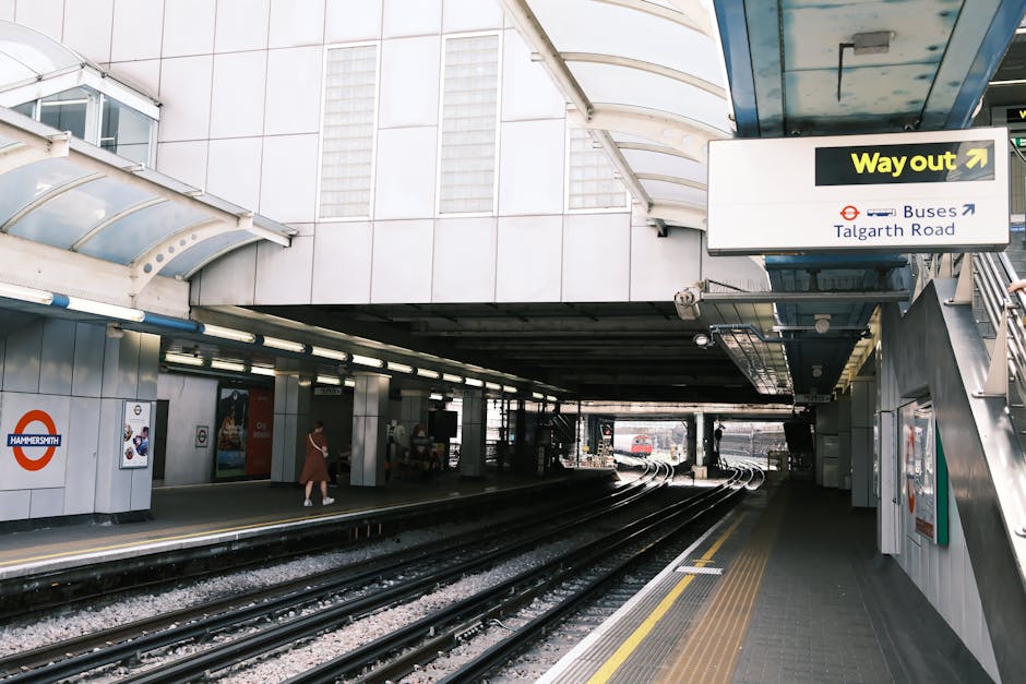 The underground station platform in Hammersmith featuring clean, polished concrete flooring with yellow tactile paving strips near the edge, stainless steel railings and supports, and a curved, transl