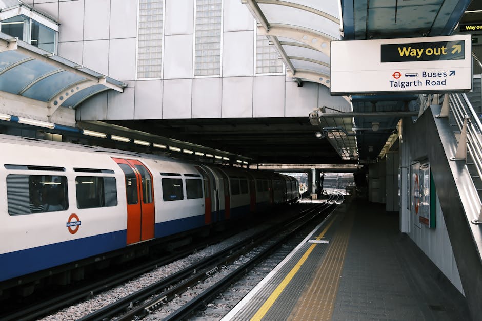 The underground station platform in Hammersmith featuring clean, polished concrete flooring with yellow tactile paving strips near the edge, stainless steel railings and supports, and a curved, transl