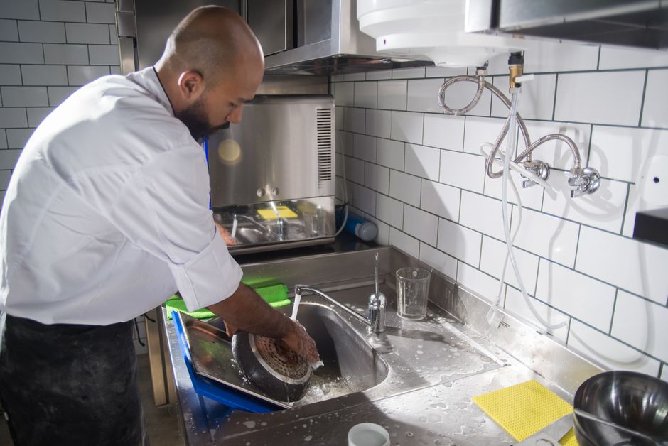 A professional cleaner wearing a white uniform is engaged in surface cleaning and sanitisation of a stainless steel kitchen sink in a commercial kitchen setting. The stainless steel sink is located ag