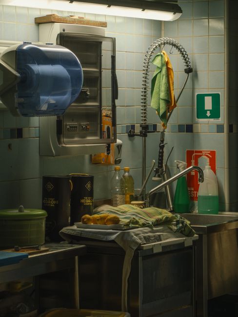 A professional cleaner wearing a white uniform is engaged in surface cleaning and sanitisation of a stainless steel kitchen sink in a commercial kitchen setting. The stainless steel sink is located ag