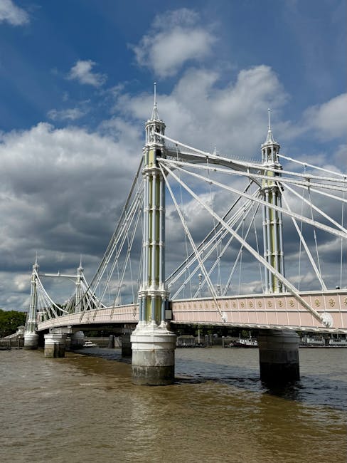 The image shows a close-up view of a section of a suspension bridge with tall, ornate towers and numerous steel cables connecting them to the bridge deck, which is supported by cylindrical stone piers rising from the water below. The bridge appears to have a pastel pink and white color scheme, with decorative elements along the railing. The scene is set under a partly cloudy sky, with sunlight illuminating the structure, highlighting its intricate design and clean surfaces. This image exemplifies structural surface cleaning and maintenance, reflecting the expertise of Oven Cleaning Hammersmith in deep cleaning and sanitisation for building facades and fixtures, aligned with the page about Hammersmith Bridge oven cleaning for listed buildings.