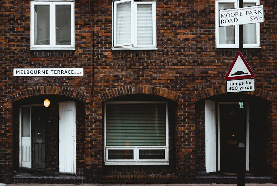 Photograph of a brick residential building at the corner of Melbourne Terrace, SW6, showing two white-framed windows on the upper floor and two front doors with arched brickwork above on the ground floor. The left door is white with a light next to it, and the right door is black with a doorbell and mail slot. The building features brick surfaces with some mortar visible and appears clean and well-maintained. Street signs indicate the location as Moore Park Road SW6 and Melbourne Terrace SW6, with one sign warning about street bumps for 450 yards. The scene is evenly lit, capturing the brick facade and signage clearly, reflecting urban residential architecture.
