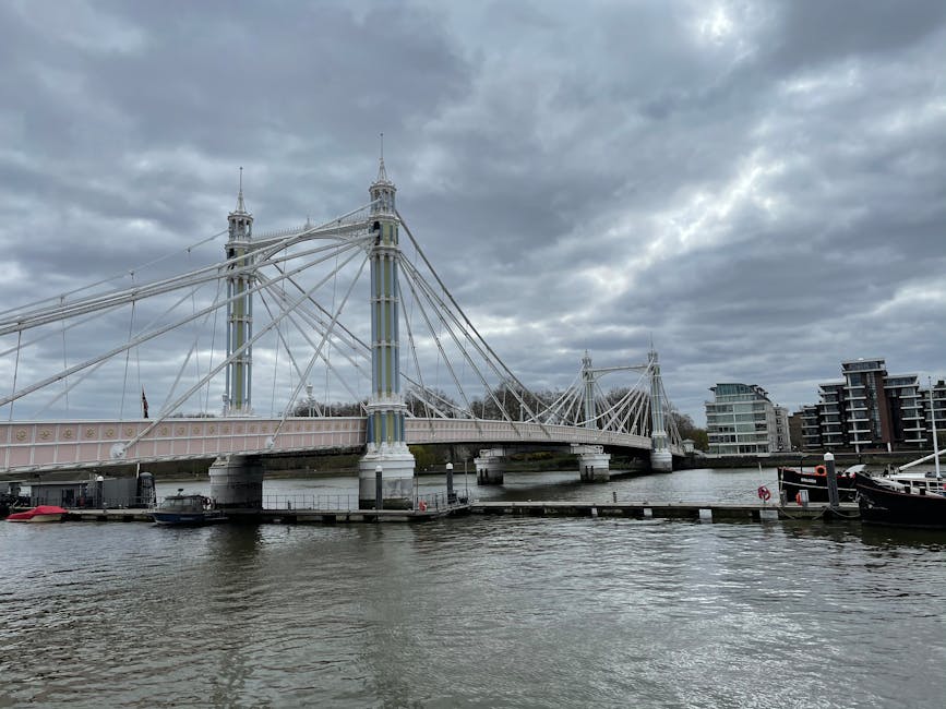 The image shows a close-up view of a section of a suspension bridge with tall, ornate towers and numerous steel cables connecting them to the bridge deck, which is supported by cylindrical stone piers rising from the water below. The bridge appears to have a pastel pink and white color scheme, with decorative elements along the railing. The scene is set under a partly cloudy sky, with sunlight illuminating the structure, highlighting its intricate design and clean surfaces. This image exemplifies structural surface cleaning and maintenance, reflecting the expertise of Oven Cleaning Hammersmith in deep cleaning and sanitisation for building facades and fixtures, aligned with the page about Hammersmith Bridge oven cleaning for listed buildings.