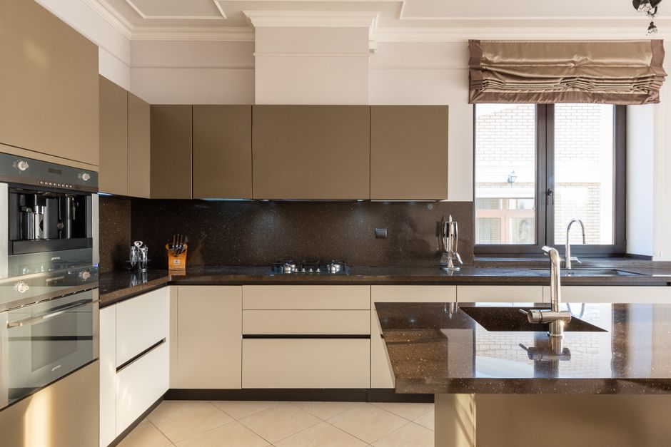 A modern kitchen featuring beige and brown cabinetry with smooth, flat surfaces, and a matching dark brown granite countertop. The kitchen includes a built-in stainless steel oven and microwave on the left, with a small jar and utensils on the countertop. A large window with a brown Roman blind allows natural light to illuminate the space, casting a bright reflection on the polished, clean granite surfaces. The sink, with a chrome faucet, is centered under the window, and the surrounding surfaces are free of dust or grime, indicating recent deep cleaning and sanitisation. The room's neutrally toned tile flooring and well-maintained surfaces highlight the thorough residential cleaning services provided by Oven Cleaning Hammersmith, reflecting hygiene and attention to detail.