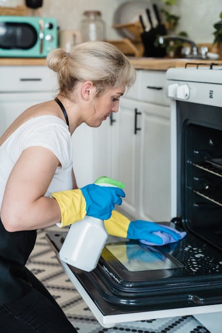 A woman with blonde hair tied in a bun, wearing a white t-shirt and yellow and blue cleaning gloves, is performing surface cleaning inside a modern kitchen. She is using a spray bottle and a cloth to clean the interior of a black oven with a glass door, which is partially open, revealing clean oven racks. The surrounding kitchen features white cabinetry with black handles, a patterned black and white rug on the floor, and various kitchen appliances, including a microwave in a light turquoise color on the countertop. The scene is well-lit with natural light, emphasizing the shiny, clean surfaces and the meticulous cleaning process. This image reflects domestic cleaning and deep sanitisation services provided by Oven Cleaning Hammersmith for residents in Ravenscourt Park.