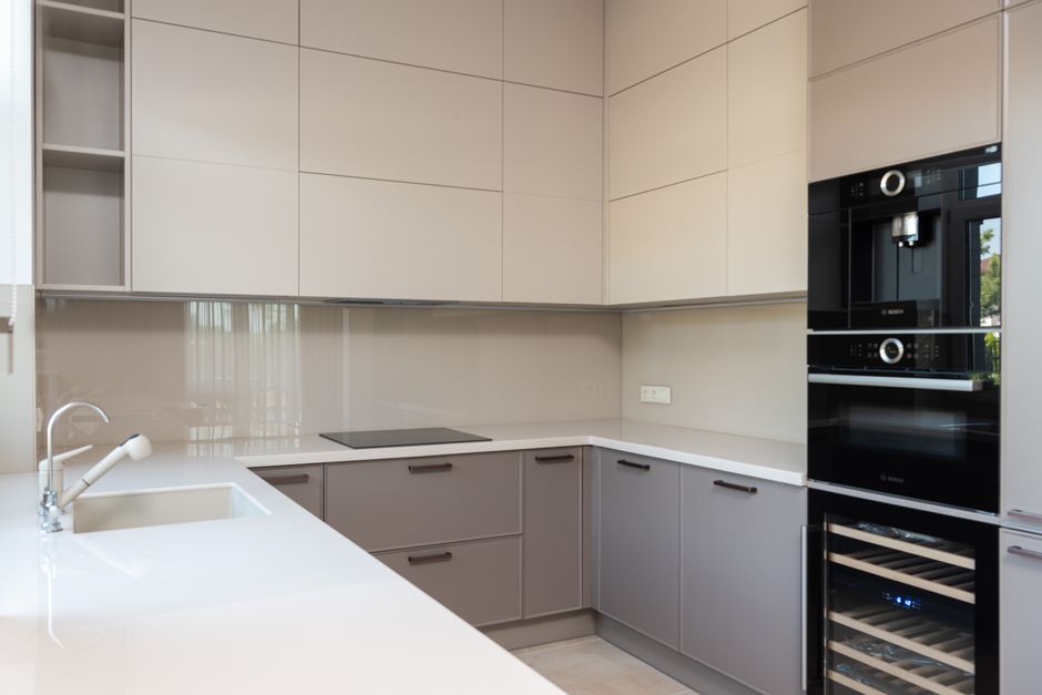 A modern kitchen featuring beige and brown cabinetry with smooth, flat surfaces, and a matching dark brown granite countertop. The kitchen includes a built-in stainless steel oven and microwave on the left, with a small jar and utensils on the countertop. A large window with a brown Roman blind allows natural light to illuminate the space, casting a bright reflection on the polished, clean granite surfaces. The sink, with a chrome faucet, is centered under the window, and the surrounding surfaces are free of dust or grime, indicating recent deep cleaning and sanitisation. The room's neutrally toned tile flooring and well-maintained surfaces highlight the thorough residential cleaning services provided by Oven Cleaning Hammersmith, reflecting hygiene and attention to detail.